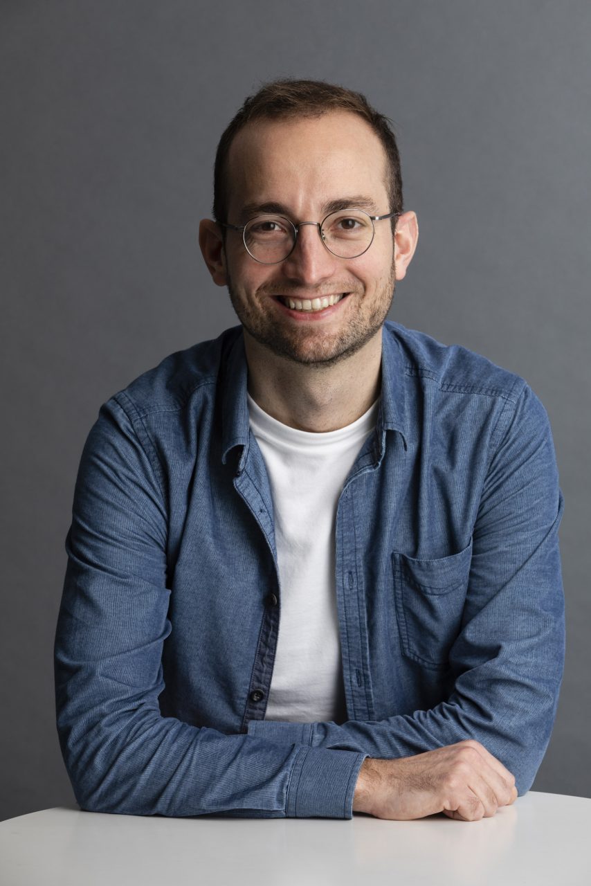 Portrait corporate d’un homme souriant avec lunettes, assis en studio sur fond gris